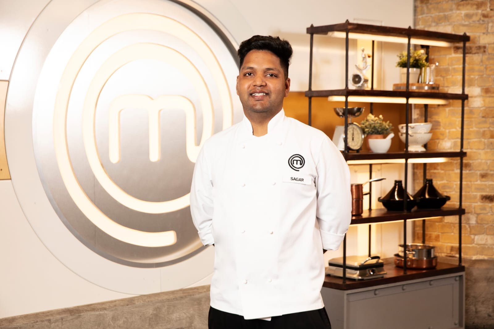Man in a white chef's jacket with hands behind back facing the camera in front of the BBC Masterchef logo on the wall behind him